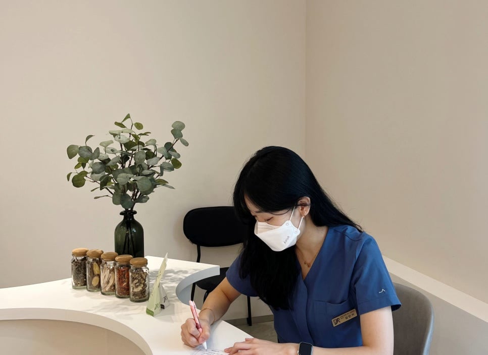 마스크를 쓴 김민지 대표원장이 약재 유리병이 놓인 상담 테이블에서 기록지를 쓰는 모습