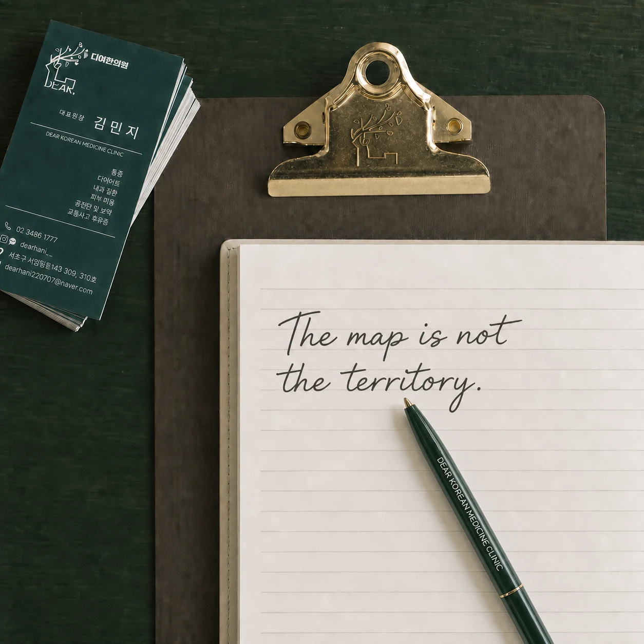 디어한의원 사슴 로고가 음각된 황동 클립과 명함, The map is not the territory.가 적힌 노트가 놓인 책상