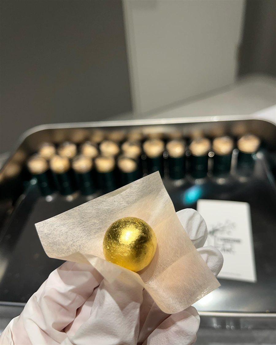 A gloved hand holding one gold-leaf Gongjindan pill, with jars on a tray behind